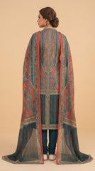 Full length front view of a model standing in a Mughal courtyard in a terracotta red luxury lawn suit with all-over paisley jaal print, plain teal shalwar, and matching teal chiffon dupatta with oversized paisley print and gold border draped over one arm