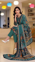 Model seated on a gold wire chair in a colourful pendant-light studio wearing a rich teal luxury lawn suit with abstract ikat floral placement print in orange, rust, and navy, V-notch neckline with embroidered orange and teal leaf motifs, multicolour ikat cuff border, plain teal cigarette trousers, and matching dark teal printed lawn dupatta with geometric chevron border pooling on the floor