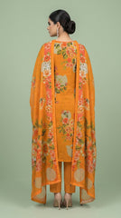 Model standing in a green-panelled studio with circular window holding an open book, wearing a marigold orange luxury lawn suit with all-over oversized watercolour peony and hibiscus floral print, V-notch neckline with fabric button placket, plain orange cigarette trousers, and matching printed lawn dupatta with coral border draped over one arm