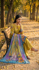 Full length side view of a model leaning against a large tree on an autumn avenue in a chartreuse olive luxury lawn suit with tonal vine and multicolour wildflower print, teal ikat cuff border, plain olive yellow palazzo trousers, and teal-to-lavender chiffon dupatta with ikat chevron border draped over the head