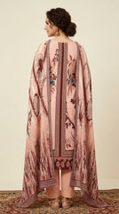 Full length front view of a model seated on a festive diwan in a blush pink luxury lawn suit with dark brown botanical floral and bird print, dark brown ikat placket panel, plain blush pink cigarette trousers, and matching printed lawn dupatta with ikat diamond border, marigold and fairy-light festive background