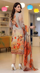 Back view of a model in a modern living room with colourful pendant lights wearing a warm beige luxury lawn suit with scattered orange-red lily and botanical floral print, embroidered cuff border, plain beige cigarette trousers, and cream-to-peach chiffon dupatta with oversized orange-red poppy and lily blooms draped over one arm