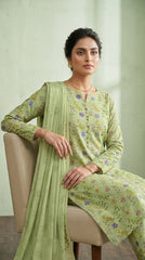 Full length front view of a model standing beside a cream armchair in a soft sage pistachio luxury lawn suit with all-over multicolour ditsy floral print, matching printed trouser, and contrast peach-gold chiffon dupatta with matching floral border draped over one shoulder, sage green wall background