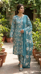 Back view of a model standing in a teal blue Pakistani luxury lawn suit with all-over scattered white cherry blossom and blush peony print, matching printed trouser, and matching sheer chiffon dupatta with oversized white peonies, warm white wall background