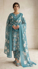 Model seated on a carved wooden Mughal bench in a warm-lit interior wearing a teal blue blossom lawn suit with matching printed trouser and matching sheer chiffon dupatta with oversized white peonies pooling on the seat, antique gold necklace and earrings