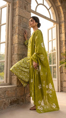 Full length side view of a model leaning against a stone archway in a golden-hour greenhouse conservatory wearing an olive chartreuse luxury lawn suit with all-over white ditsy floral print, matching printed trouser, and matching chiffon dupatta with oversized white hibiscus print draped over one shoulder